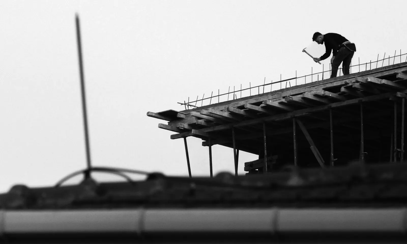 A lone worker on the edge of a concrete roof deck, bent over with a hammer against an open pale sky.