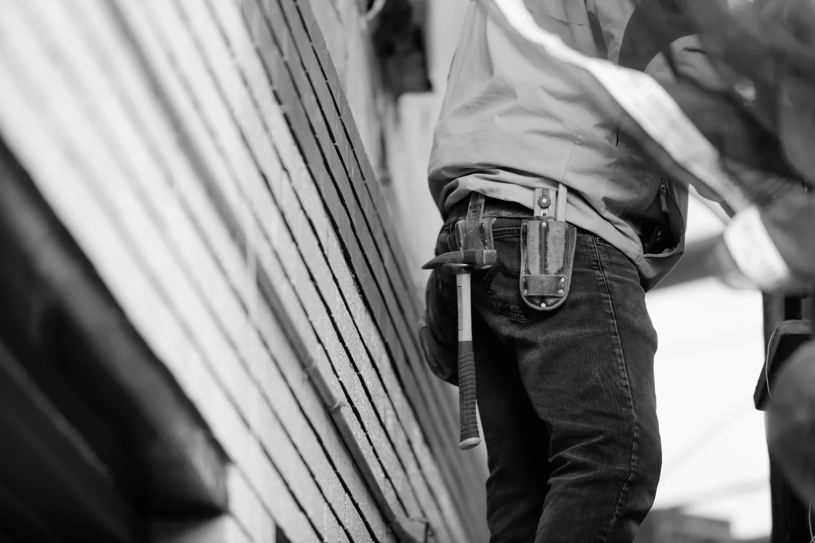 A tradesman at work beside a brick wall, seen from the waist, a claw hammer and a steel pouch hanging from his leather belt.