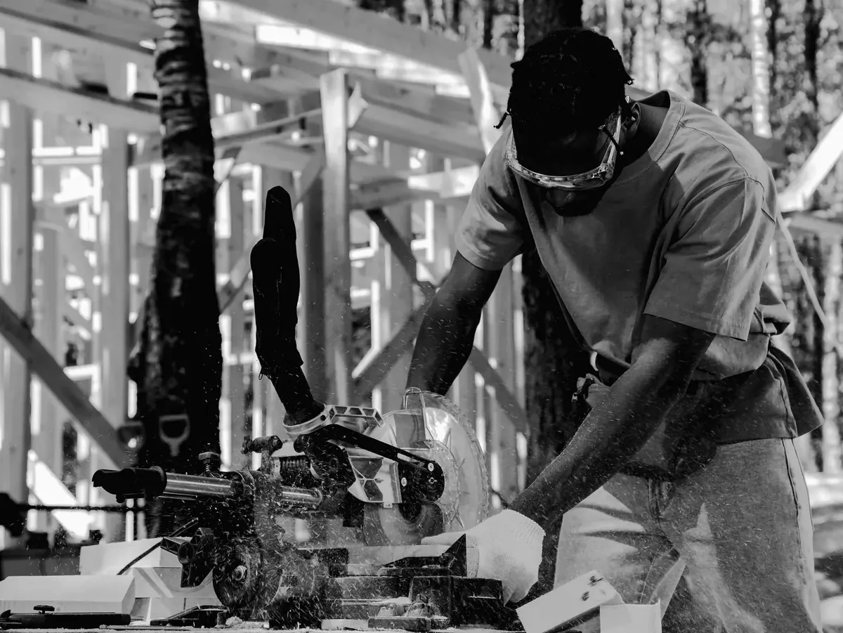 A carpenter running a board through a mitre saw on a timber frame site, sawdust in the air.