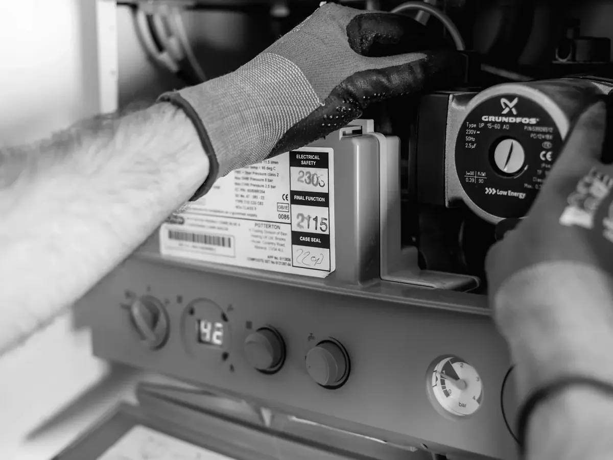 A plumber’s gloved hands adjusting the circulating pump inside an open domestic boiler, the control dials below it.